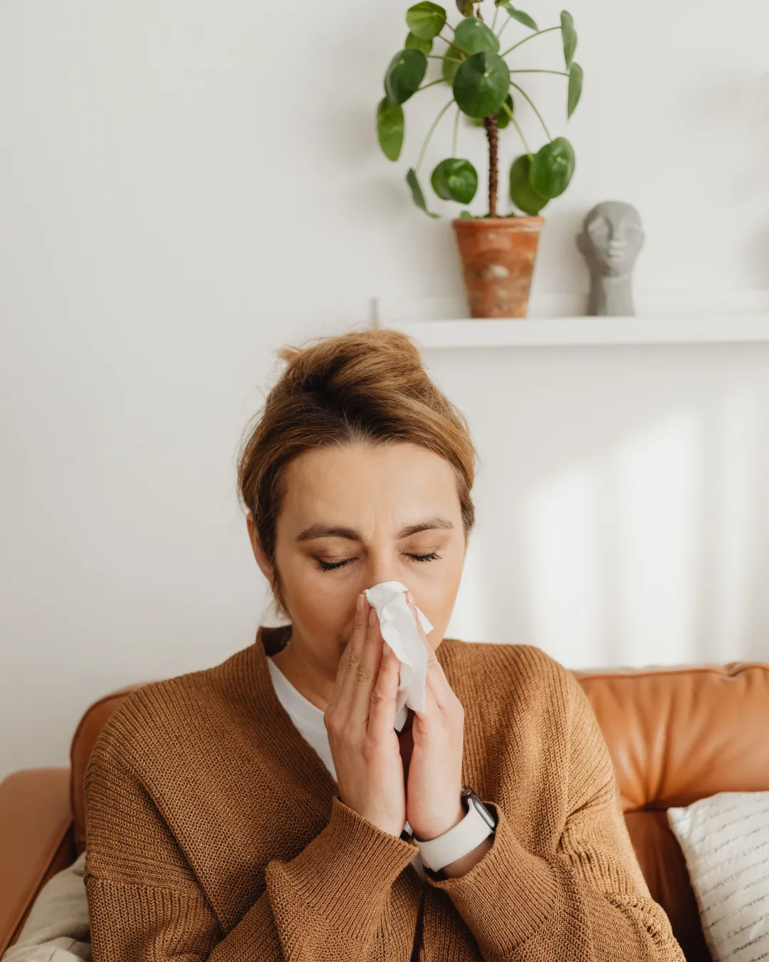 A woman sitting on a sofa at home, eyes closed, holding a tissue to her nose
